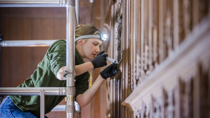 Close up of young female conservator on scaffold tower wearing head torch and black gloves 'injecting' repair treatment with syringe into damaged wood in The Vyne's Tudor Oak Gallery